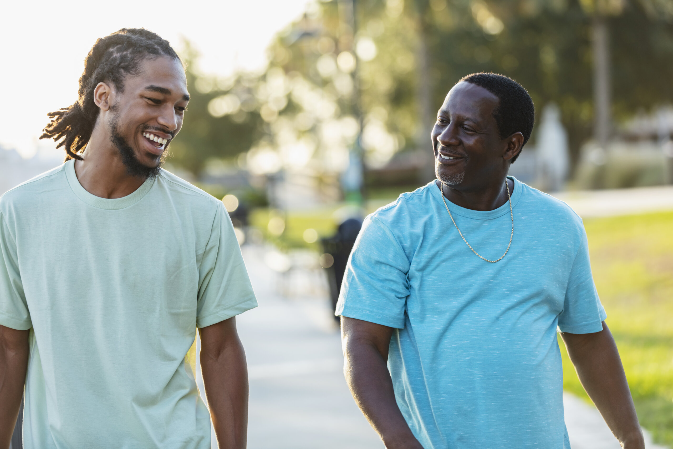 An African-American man walking and conversing with his adult son in a city park. They are smiling, looking toward each other. The young man is in his 20s, and his father is in his 50s.