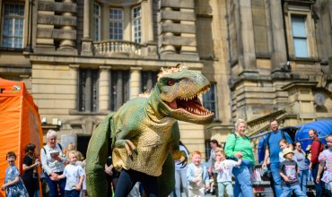 A large dinosaur costume interacts with a crowd of children and adults in front of a large stone building.