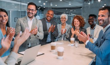 Business people clapping hands during business presentation in board room. Group of corporate business persons on a business meeting in modern office. Cheerful team sitting on conference table in board room and clapping with hands after business presentation. Group of happy business people standing in conference hall and applauding.