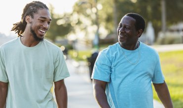 An African-American man walking and conversing with his adult son in a city park. They are smiling, looking toward each other. The young man is in his 20s, and his father is in his 50s.
