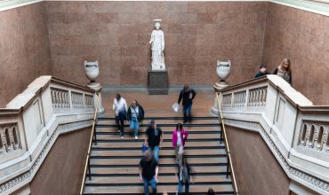 A dynamic scene of tourists walking down a grand staircase with a statue centerpiece in the British Museum